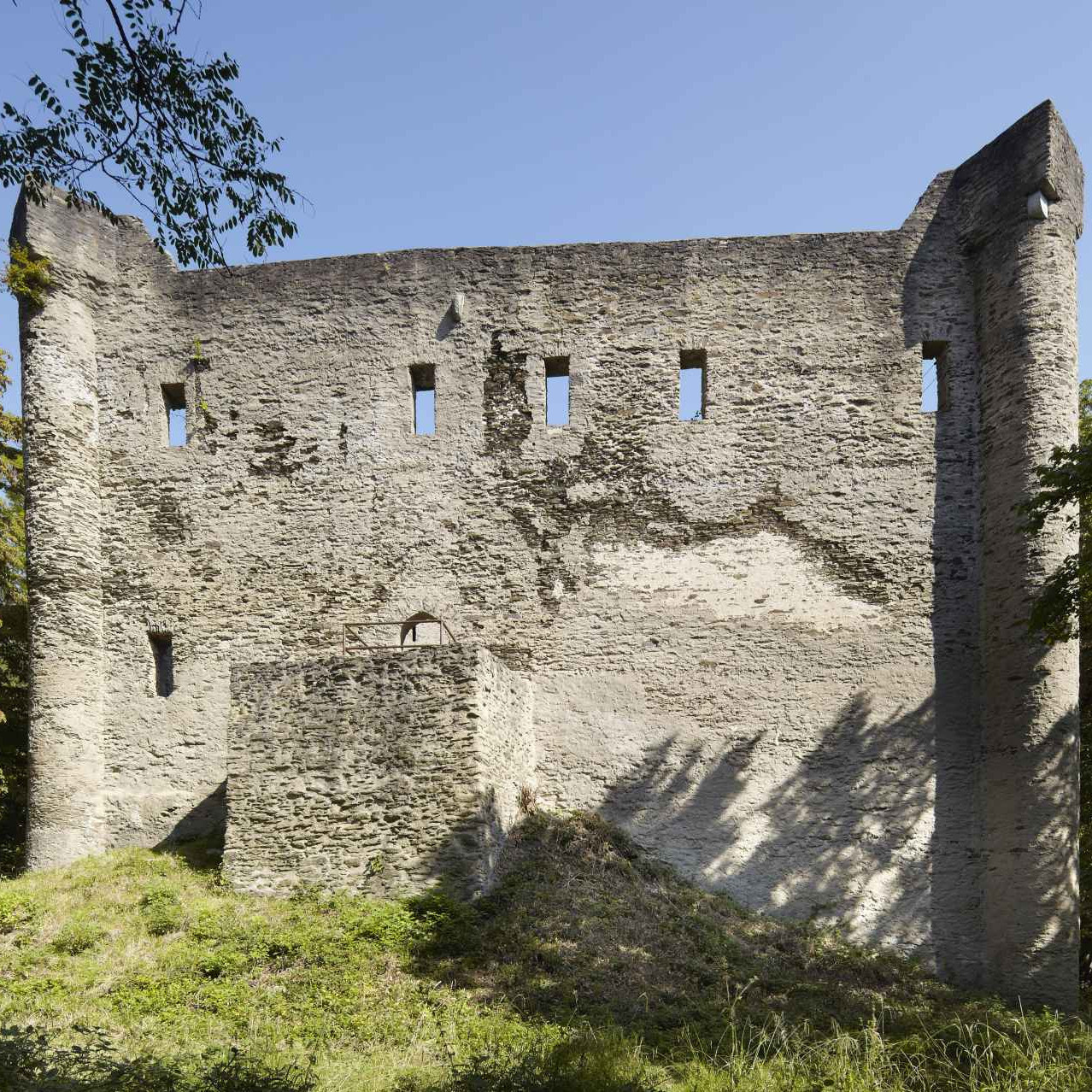 Mauer mit Vorbau hinter einem bewachsenen Hügel zwischen Bäumen