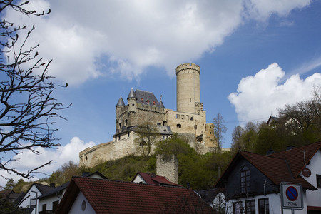 Ansicht der Burg Schwalbach, Bergfried rechts vor blauem Himmel mit Wolken, links im Vordergrund und rechts Bäume, ein Wohnhaus.