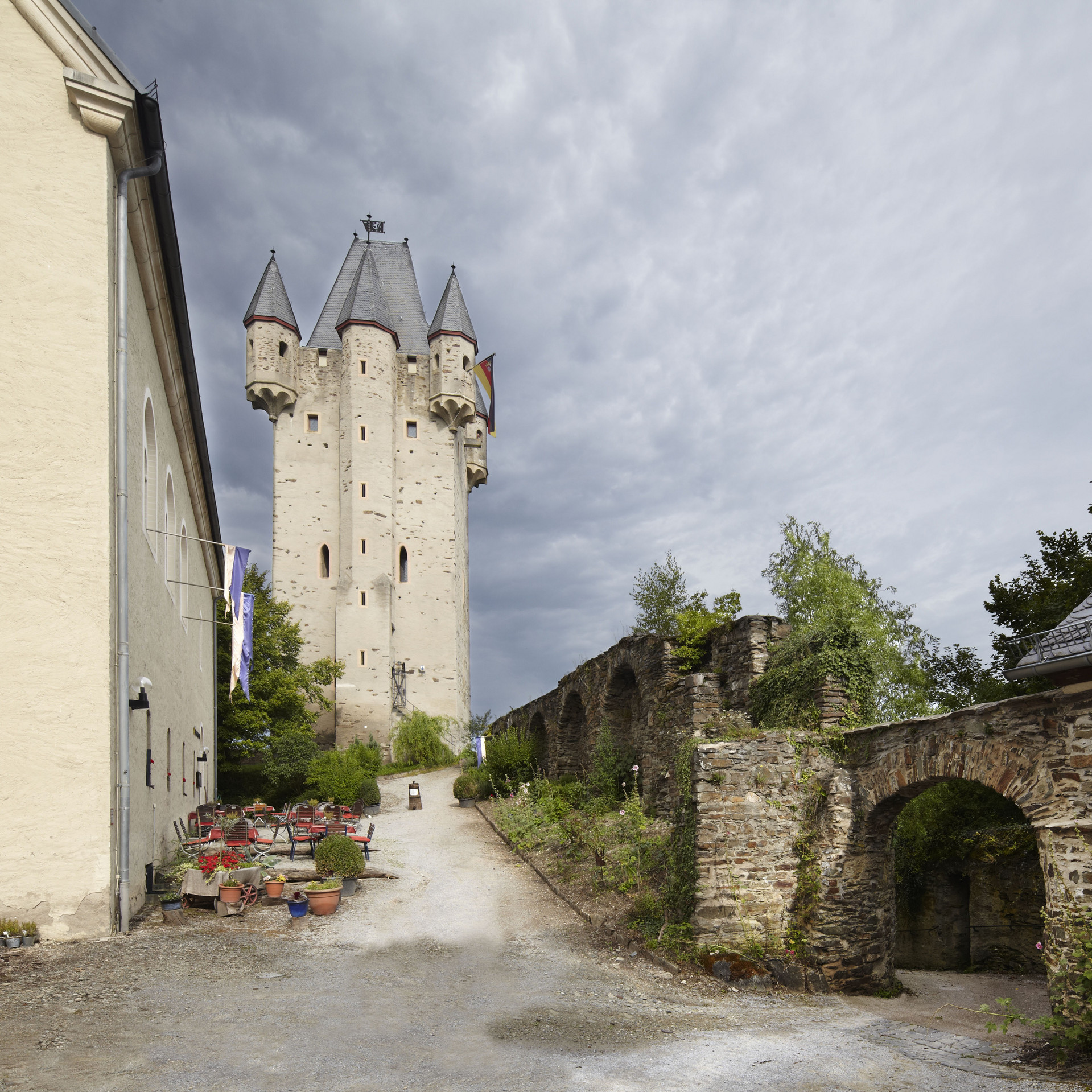 Bergfried mit Zuweg, Haus mit Tischen und Stühlen, Blumenkübeln und Korb links, Mauer mit Durchgang und Haus links