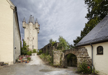 Bergfried mit Zuweg, Haus mit Tischen und Stühlen, Blumenkübeln und Korb links, Mauer mit Durchgang und Haus links