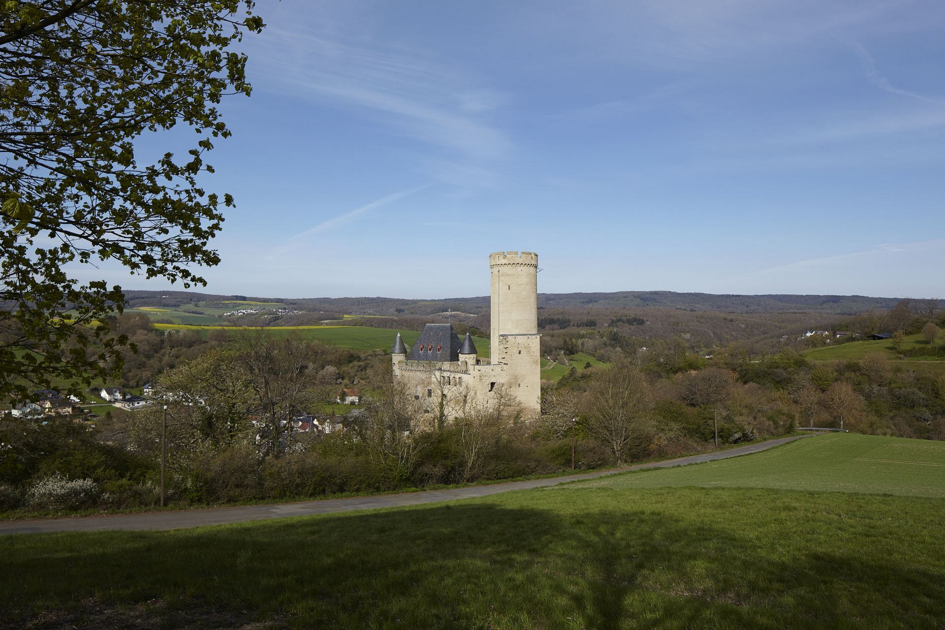 Burg Schwalbach Burg Schwalbach umgeben von Wiesen und bewaldeten Hügeln, im Hintergrund Dörfer, im Vordergrund eine Straße und Laub am linken Bildrand