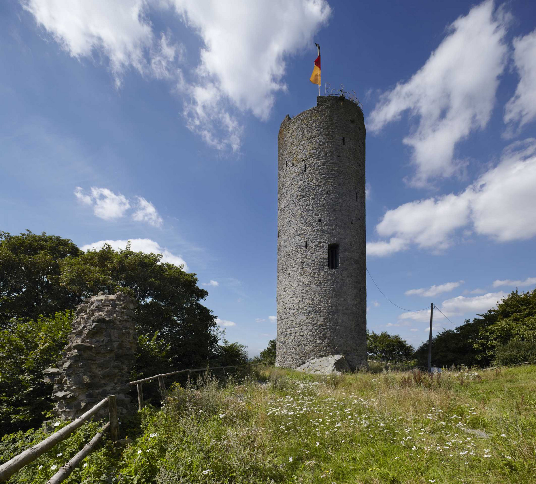 Blick auf den Bergfried, davor Wiese, links Mauerreste, im Hintergrund Bäume